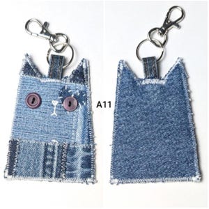 May include: A cat-shaped keychain made from blue denim fabric patches with white stitched edges. The front features two purple button eyes and white embroidered details. A silver metal lobster clasp and ring are attached to the top. Text A11 is visible.