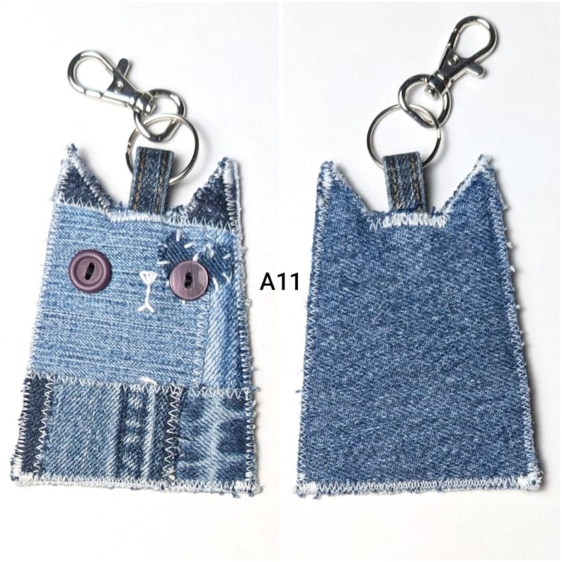 May include: A cat-shaped keychain made from blue denim fabric patches with white stitched edges. The front features two purple button eyes and white embroidered details. A silver metal lobster clasp and ring are attached to the top. Text A11 is visible.