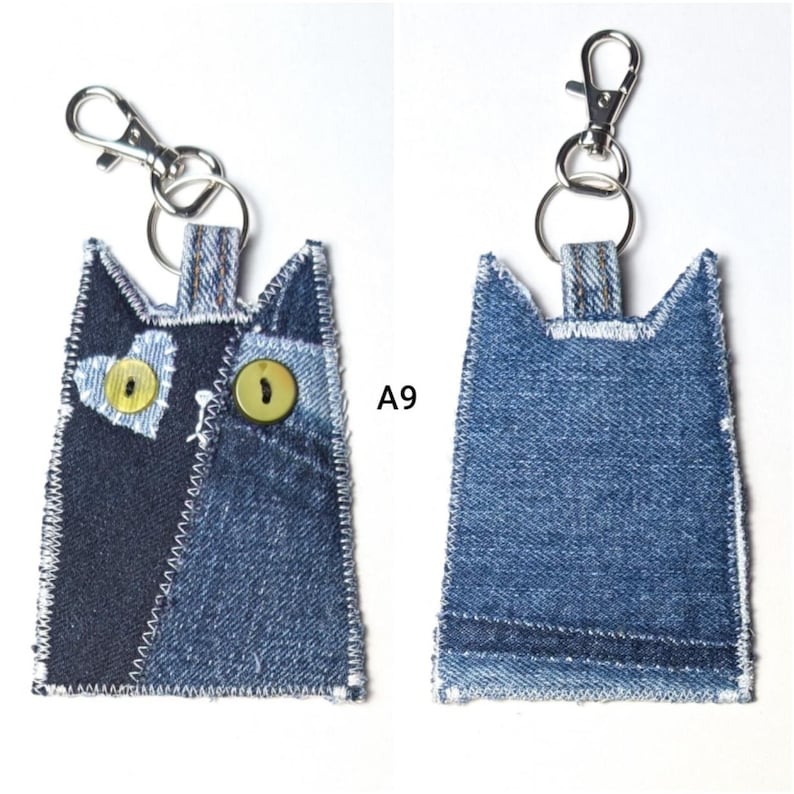 May include: A cat-shaped keychain made from blue denim fabric with white stitched edges. The front features a heart-shaped denim patch, two round yellow buttons for eyes, and a metal lobster clasp. The back is plain blue denim. Text A9 is visible.