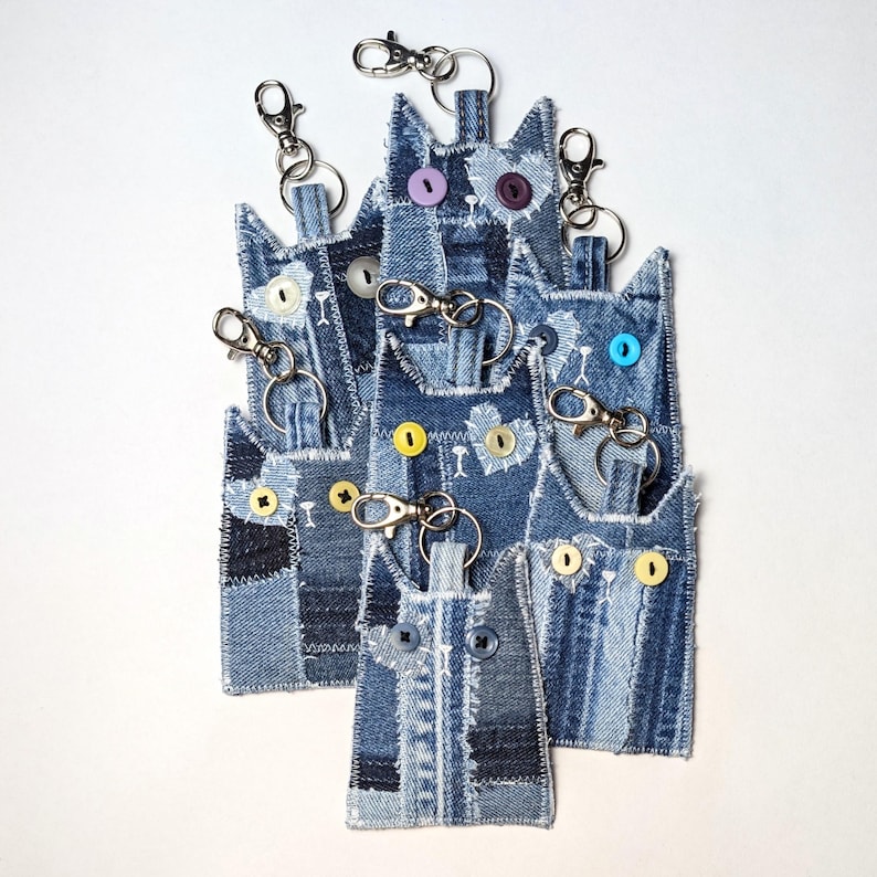 May include: A collection of cat-shaped keychains crafted from various shades of blue denim fabric. Each keychain features stitched heart details, frayed edges, and colorful button eyes. Silver metal lobster claw clasps are attached to the top.