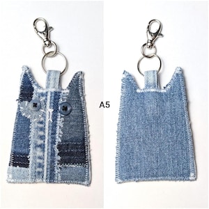 May include: A cat-shaped keychain made from blue denim fabric with frayed edges. The front features two dark blue buttons as eyes and a small heart-shaped patch. A silver metal lobster clasp and ring are attached to the top. The text A5 is visible.