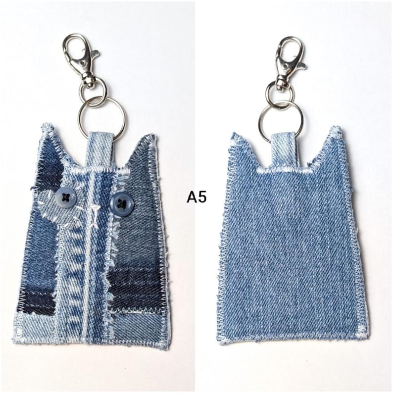 May include: A cat-shaped keychain made from blue denim fabric with frayed edges. The front features two dark blue buttons as eyes and a small heart-shaped patch. A silver metal lobster clasp and ring are attached to the top. The text A5 is visible.