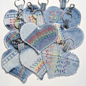 May include: A collection of denim heart-shaped keychains. Each keychain features a silver-colored clasp and a denim heart with colorful embroidered details. Some hearts have rainbow stitching, while others have geometric patterns.