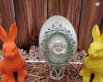 Puede incluir: Una pieza decorativa ovalada y ornamentada con un marco plateado, detalles verdes y una pequeña figura de querubín. A los lados, figuras de conejos naranja y amarillo, sobre un fondo de tablones de madera y paja.