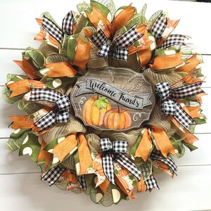 May include: A fall-themed wreath with orange, green, and black and white checkered ribbon. The wreath has a wooden sign that says "Welcome Friends".