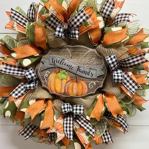 May include: A fall-themed wreath with orange, green, and black and white checkered ribbon. The wreath features a wooden sign with the text "Welcome Friends" and pumpkins.