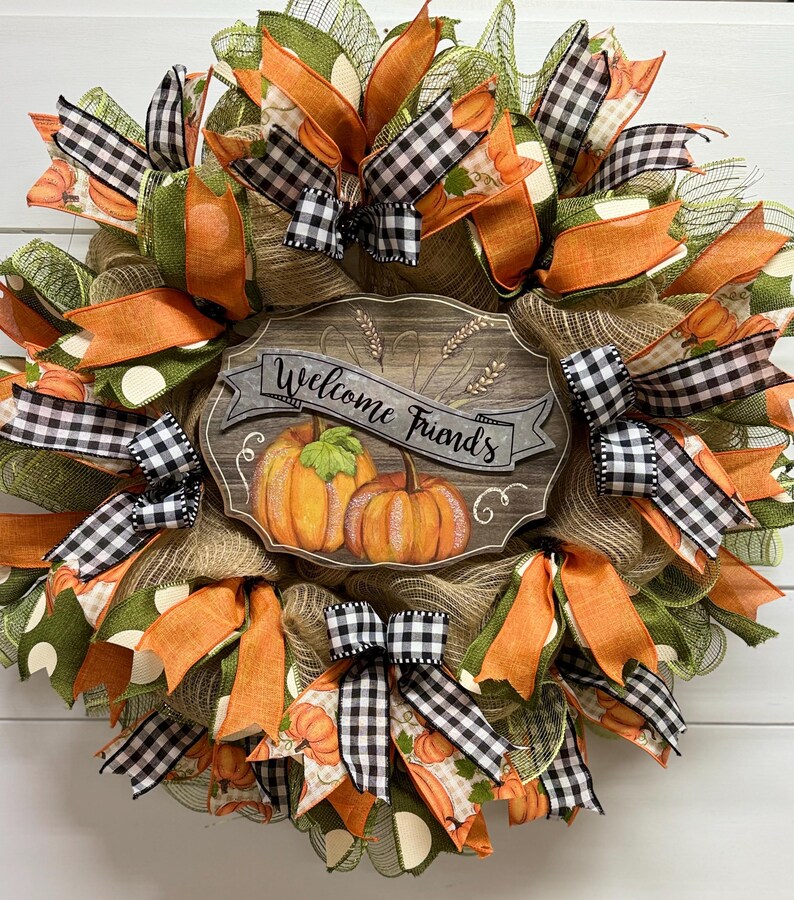 May include: A fall-themed wreath with orange, green, and black and white checkered ribbon. The wreath features a wooden sign with the text "Welcome Friends" and pumpkins.