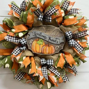 May include: A fall-themed wreath with orange, green, and black and white checkered ribbon. The wreath features a wooden sign that says "Welcome Friends" with a painted pumpkin design.