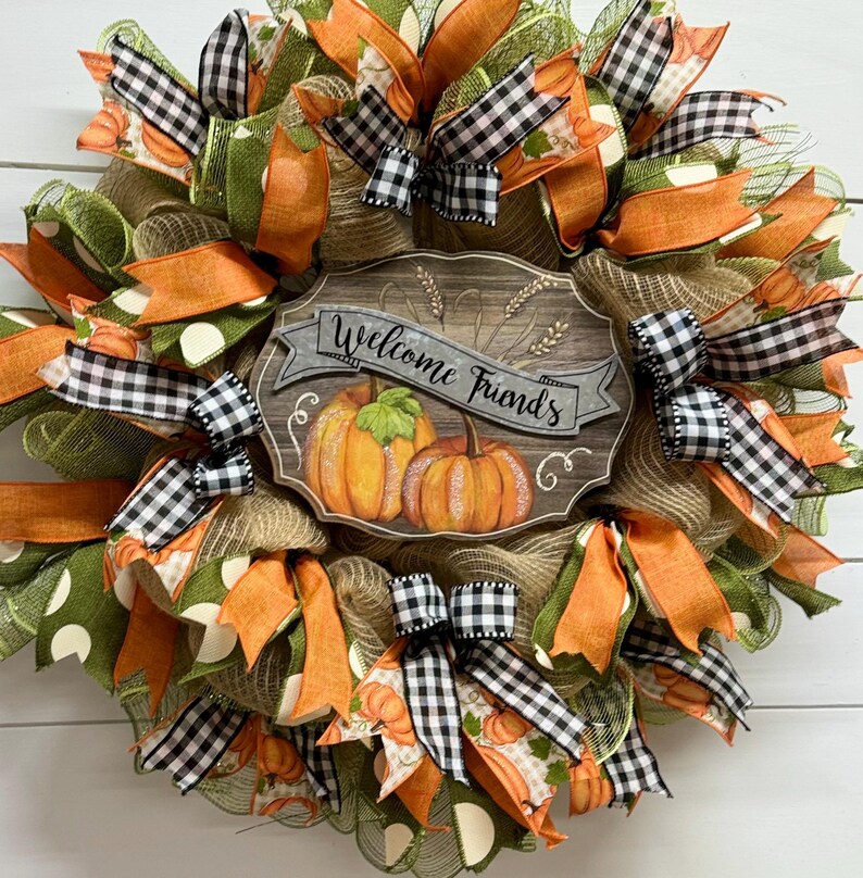 May include: A fall-themed wreath with orange, green, and black and white checkered ribbon. The wreath features a wooden sign that says "Welcome Friends" with a painted pumpkin design.