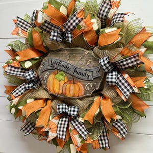 May include: A green, orange, and black fall wreath with a wooden sign that says "Welcome Friends". The wreath is decorated with pumpkins, plaid ribbon, and polka dot ribbon.