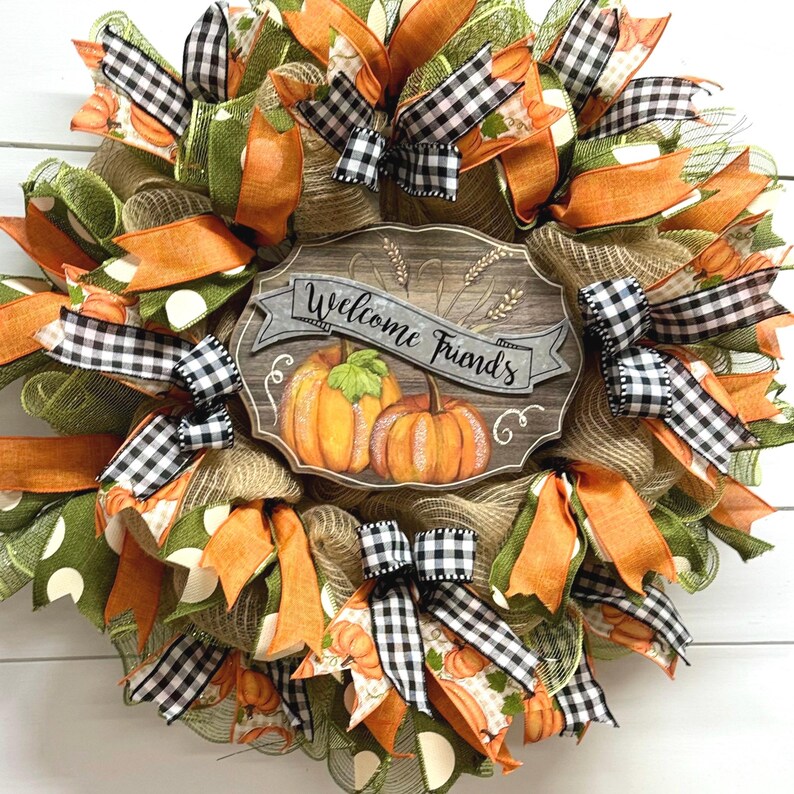 May include: A fall-themed wreath with orange, green, and black and white checkered ribbon. The wreath has a wooden sign that says "Welcome Friends" with a painted pumpkin design.