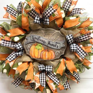 May include: A fall-themed wreath with a burlap base, orange and green mesh, black and white checkered ribbon, and a wooden sign that reads "Welcome Friends". The sign features a painted image of two pumpkins.