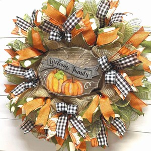 May include: A green, orange, and black fall wreath with a wooden sign that says "Welcome Friends". The wreath is decorated with pumpkins, plaid ribbon, and polka dot mesh.
