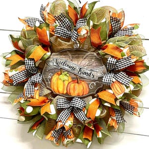 May include: A fall-themed wreath with orange, green, and black and white checkered ribbon. The wreath features a wooden sign with the text "Welcome Friends" and a painted image of pumpkins.