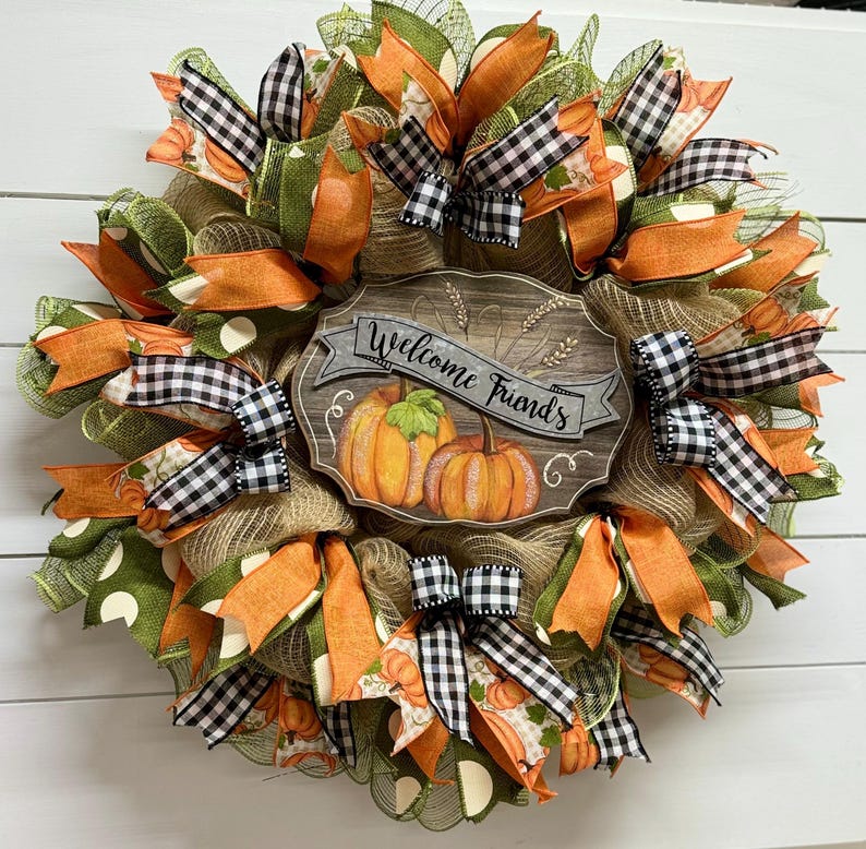 May include: A fall-themed wreath with orange, green, and black and white checkered ribbon. The wreath has a wooden sign that says "Welcome Friends".