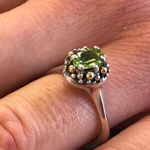 May include: A silver ring featuring a green gemstone set in a gold and silver bezel. The ring is decorated with small gold beads around the bezel.