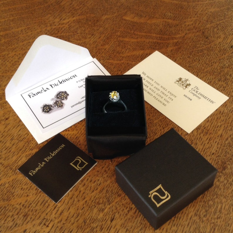 May include: A silver ring with a yellow gemstone in a black ring box. The box has a gold logo with the letter "P" on it. The ring is in a white envelope with a business card that says "Pamela Dickinson".