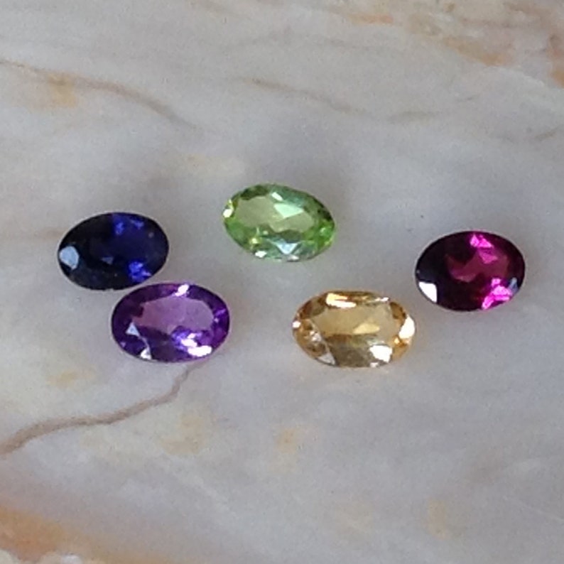May include: Five oval-shaped gemstones in various colours: purple, blue, green, yellow, and red. The gemstones are arranged in a semi-circle on a white marble surface.