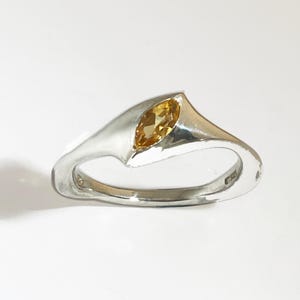 Peut inclure: Bague en argent avec une pierre précieuse jaune doré de forme marquise. La bague présente un design asymétrique moderne avec une finition polie. La pierre est sertie dans un sertissage à griffes, et le corps est lisse et incurvé.