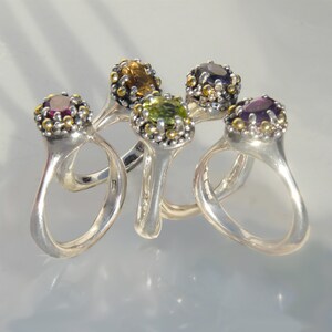 May include: Five silver rings with different coloured gemstones. The rings are stacked together and the gemstones are visible. The gemstones are purple, green, yellow, and pink.