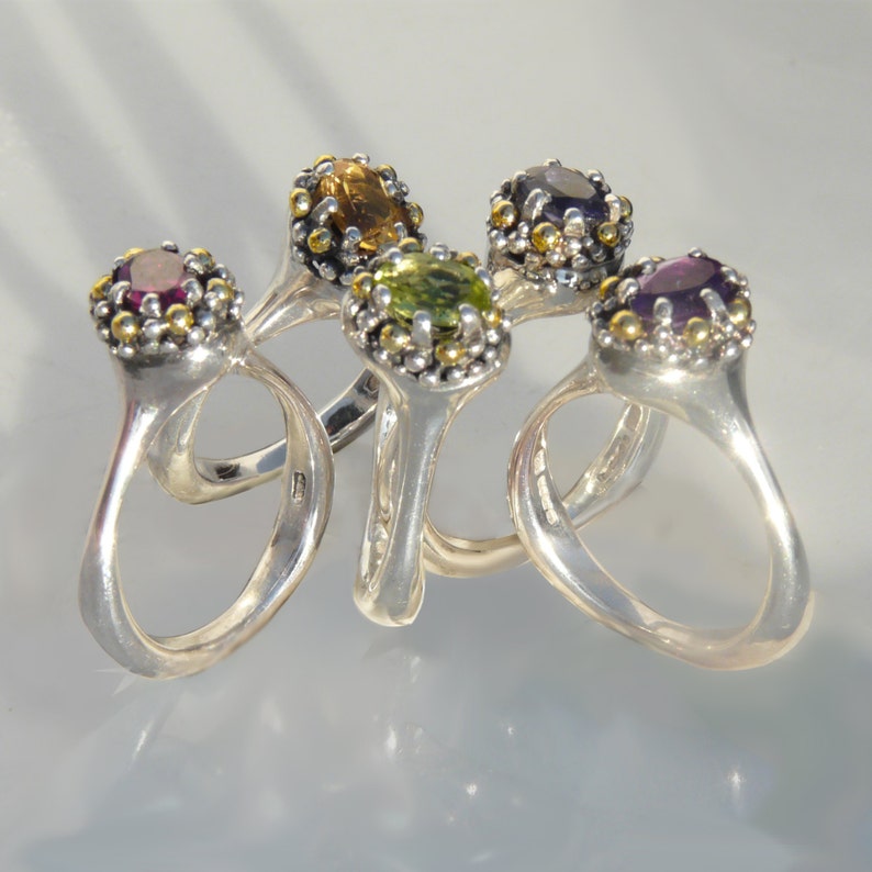 May include: Five silver rings with different coloured gemstones. The rings are stacked together and the gemstones are visible. The gemstones are purple, green, yellow, and pink.