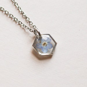 May include: A silver chain necklace with a hexagonal pendant featuring a pressed blue forget-me-not flower encased in clear resin.