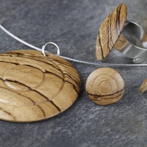 May include: A set of wooden jewelry featuring a pendant, a ring, and earrings. The pendant is a round disc with concentric circles of wood grain. The ring is a silver band with a wooden top. The earrings are small round discs with wood grain.
