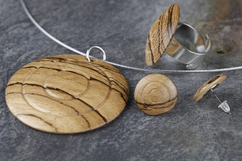 May include: A set of wooden jewelry featuring a pendant, a ring, and earrings. The pendant is a round disc with concentric circles of wood grain. The ring is a silver band with a wooden top. The earrings are small round discs with wood grain.