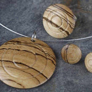 May include: A set of wooden jewelry featuring a pendant, earrings, and a ring. The pieces are made of wood with a natural grain pattern and a dark brown color. The pendant is a large circle with a smaller circle in the center. The earrings are small circles, and the ring is a thin band with a small circle on top.