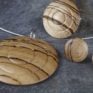 May include: A set of wooden jewelry featuring a pendant and earrings. The pendant is a large circle with concentric rings of wood grain. The earrings are smaller circles with a similar wood grain pattern.