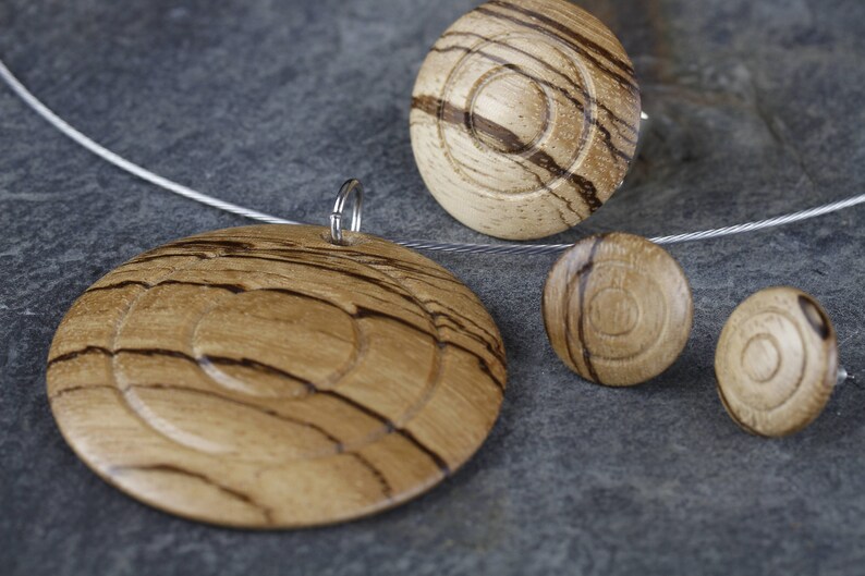 May include: A set of wooden jewelry featuring a pendant and earrings. The pendant is a large circle with concentric rings of wood grain. The earrings are smaller circles with a similar wood grain pattern.