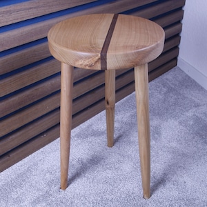 Könnte beinhalten: Ein kleiner, runder Holzschemel mit drei Beinen. Der Sitz des Hockers ist hellbraun mit einem dunkelbraunen Streifen in der Mitte. Die Beine sind in einem helleren Braunton gehalten. Der Hocker ist aus Holz und hat eine natürliche Oberfläche.