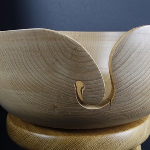 Könnte beinhalten: Eine helle Holz-Garn-Schale mit einem einzigartigen Design mit zwei ausgeschnittenen Schlaufen an der Seite. Die Schale steht auf einem Holzhocker.