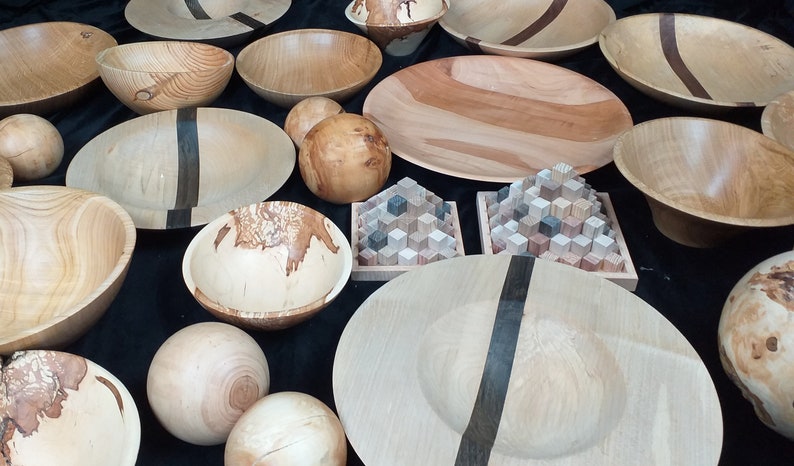 May include: A collection of wooden bowls and spheres, some with dark wood accents, displayed on a black surface. The bowls are various sizes and shapes, with some featuring a natural wood grain pattern.