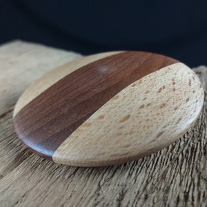 May include: A round wooden coaster with a light brown and dark brown wood grain pattern. The coaster is made of two different types of wood.