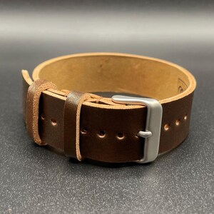 May include: A brown leather watch strap with a silver-colored buckle. The strap is thick and has several holes for adjustment. The leather appears to be of high quality, with a natural grain. The watch band is designed to be durable and stylish.