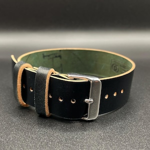 May include: A black leather watch strap with a silver buckle. The strap has a light brown edge and a green interior. The strap is shown on a dark background.