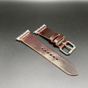 May include: A brown leather watch band with silver-colored connectors and buckle. The band is made of a smooth, dark brown leather and features several holes for adjustment. The buckle is a simple, rectangular design.