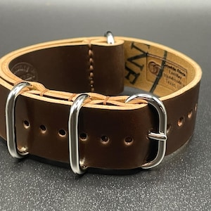May include: A brown leather watch strap with silver-colored hardware. The strap has a light-colored interior and is marked with the text "NVE" and "Handmade Texas, USA". The strap features multiple adjustment holes.