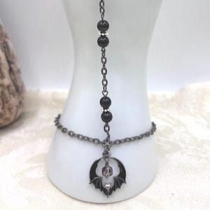 May include: A gunmetal slave bracelet featuring a delicate chain connected to a ring loop. The design includes four smooth black beads accented with small silver-toned spacers and a central circular pendant depicting a bat with spread wings.