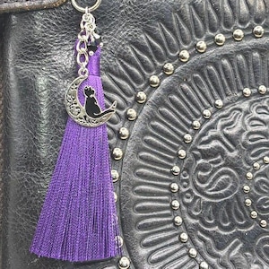 May include: A purple tassel charm with a silver cat-on-the-moon pendant and black beads, attached to a silver clasp. The charm hangs from a black leather bag with a decorative embossed design.