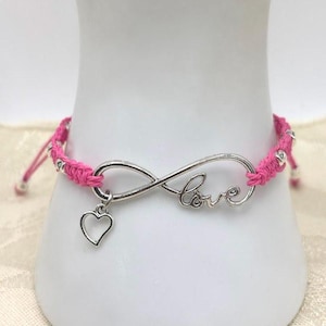 May include: Pink woven bracelet featuring a silver infinity symbol with the word "love" and a heart charm. The bracelet has silver beads and adjustable ties.