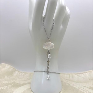 May include: A silver-toned hand chain bracelet featuring a cloud-shaped pendant and dangling crystals. The cloud pendant is white and textured, with a delicate chain connecting it to the bracelet. The bracelet is displayed on a white mannequin hand.