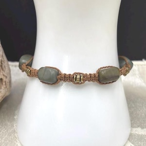 May include: A handmade bracelet featuring olive-green stone beads and small brass accents. The beads are strung on a brown macrame cord. The bracelet is displayed on a white, cylindrical object, showcasing the intricate knotting and natural stone elements.
