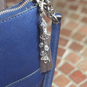 May include: A blue handbag with a silver zipper and strap. A silver charm with a tassel, clear beads, and umbrella charms hangs from the bag's handle. The background is a brick wall.