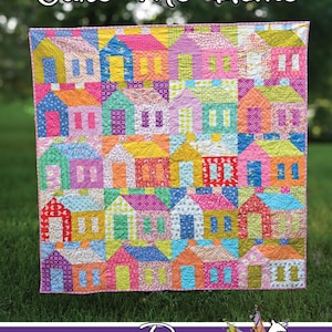 May include: A quilt featuring a grid of colorful house blocks in various patterns and shades of pink, yellow, blue, orange, and green. The text Cake Me Home appears at the top, and Running Doe Quilts For Villa Rosa Designs is at the bottom.