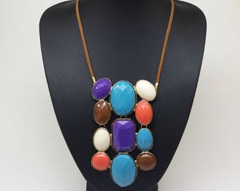Op de afbeelding: Een opvallende ketting met een hanger van kleurrijke, onregelmatig gevormde edelstenen in paars, blauw, bruin, wit en oranje. De hanger hangt aan een bruine koordketting en wordt tentoongesteld op een zwarte mannequin.