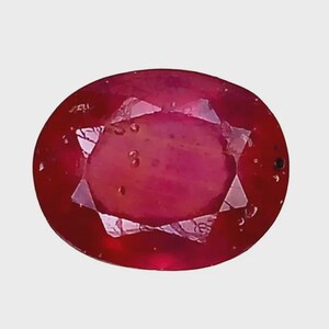 May include: An oval-cut ruby gemstone with a deep red color and a faceted surface. The stone displays internal inclusions and a polished finish, reflecting light across its multiple triangular and trapezoidal facets throughout the entire surface.