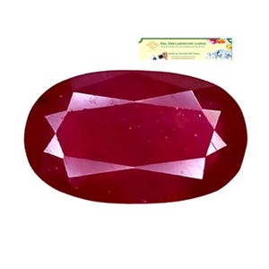 May include: An oval-cut ruby gemstone with a deep red color and a faceted surface. The stone features a central rectangular facet surrounded by triangular and trapezoidal facets. A small rectangular certificate card labeled EGL GEM LABORATORY JAPAN is visible.
