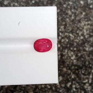 以下が含まれることがあります： 鮮やかな赤色で楕円形にカットされたルビーの宝石。ファセット加工された表面を持ち、滑らかで白い長方形のディスプレイブロックの端に配置されており、その透明度と光の反射特性を際立たせています。